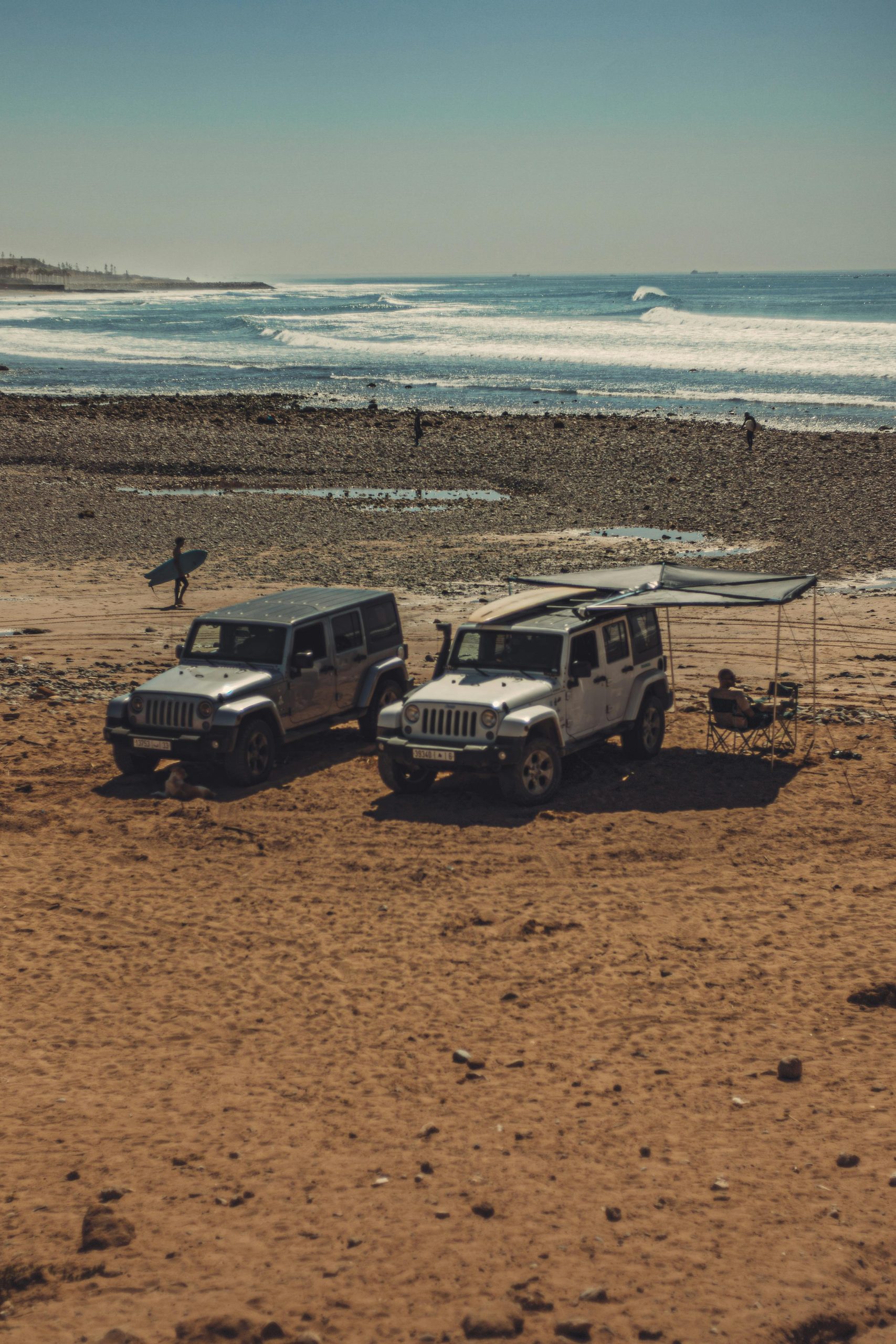 SUVs parked on a sandy beach with surfers in the distance enjoying the waves.